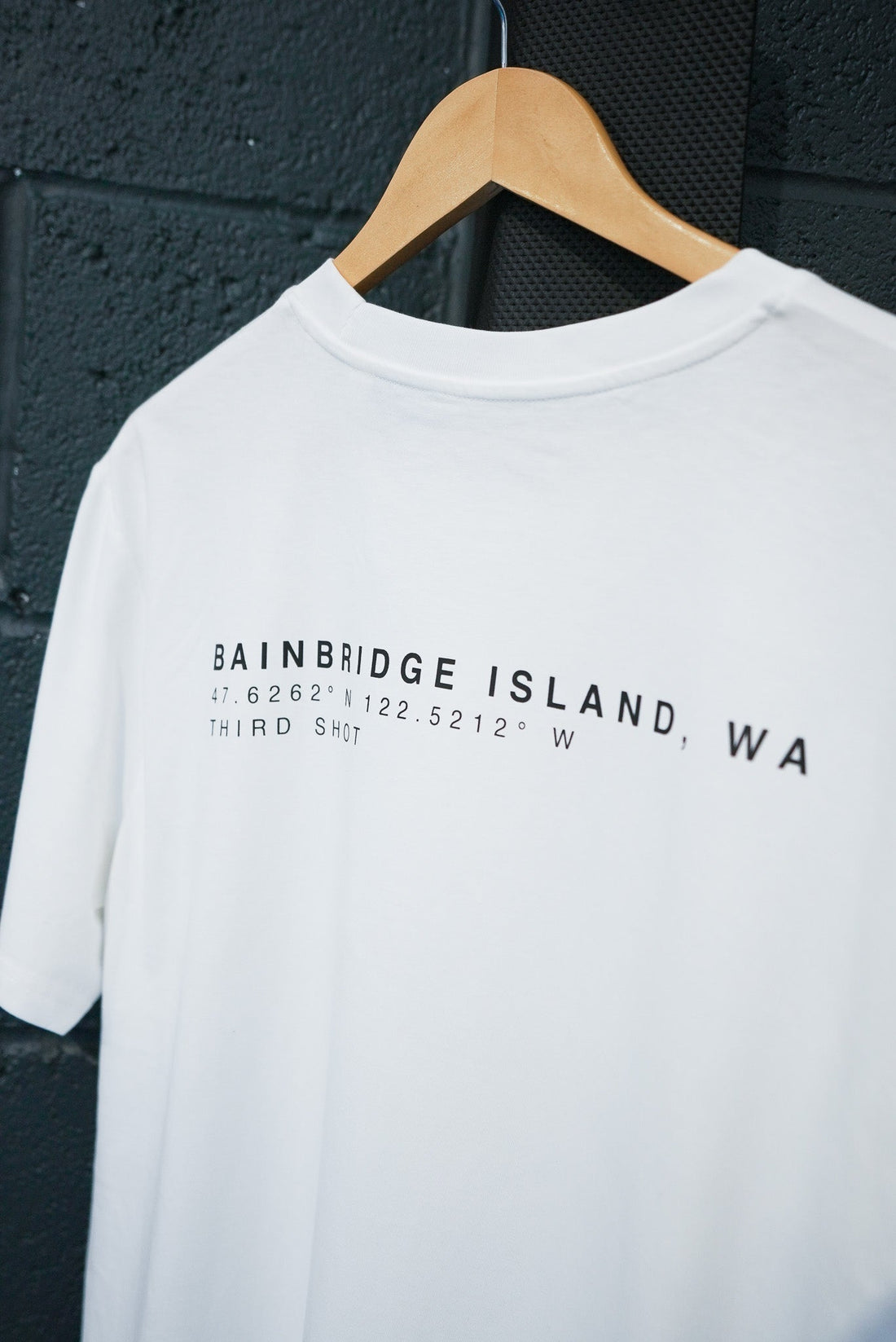 White T-shirt on wooden hanger with black text 'Bainbridge Island, WA' and coordinates on black wall background