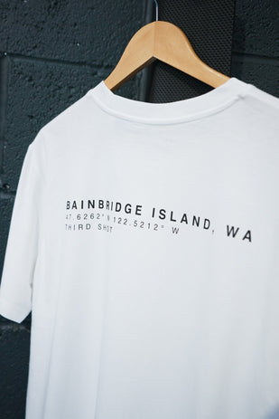 White T-shirt on wooden hanger with black text 'Bainbridge Island, WA' and coordinates on black wall background