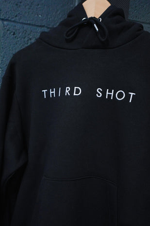 Black hoodie with white embroidered text "THIRD SHOT" on chest against dark textured wall