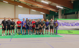 Group of people holding pickleball paddles on indoor court with Barbados banners behind them
