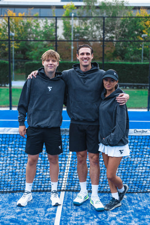 Three young adults in black athletic hoodies posing on a blue padel tennis court with net and greenery background