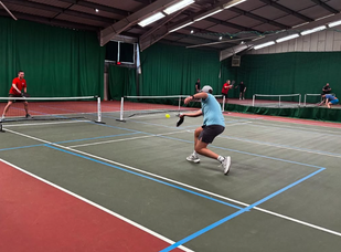 Indoor pickleball game with players in active poses on green court with blue lines and green curtains