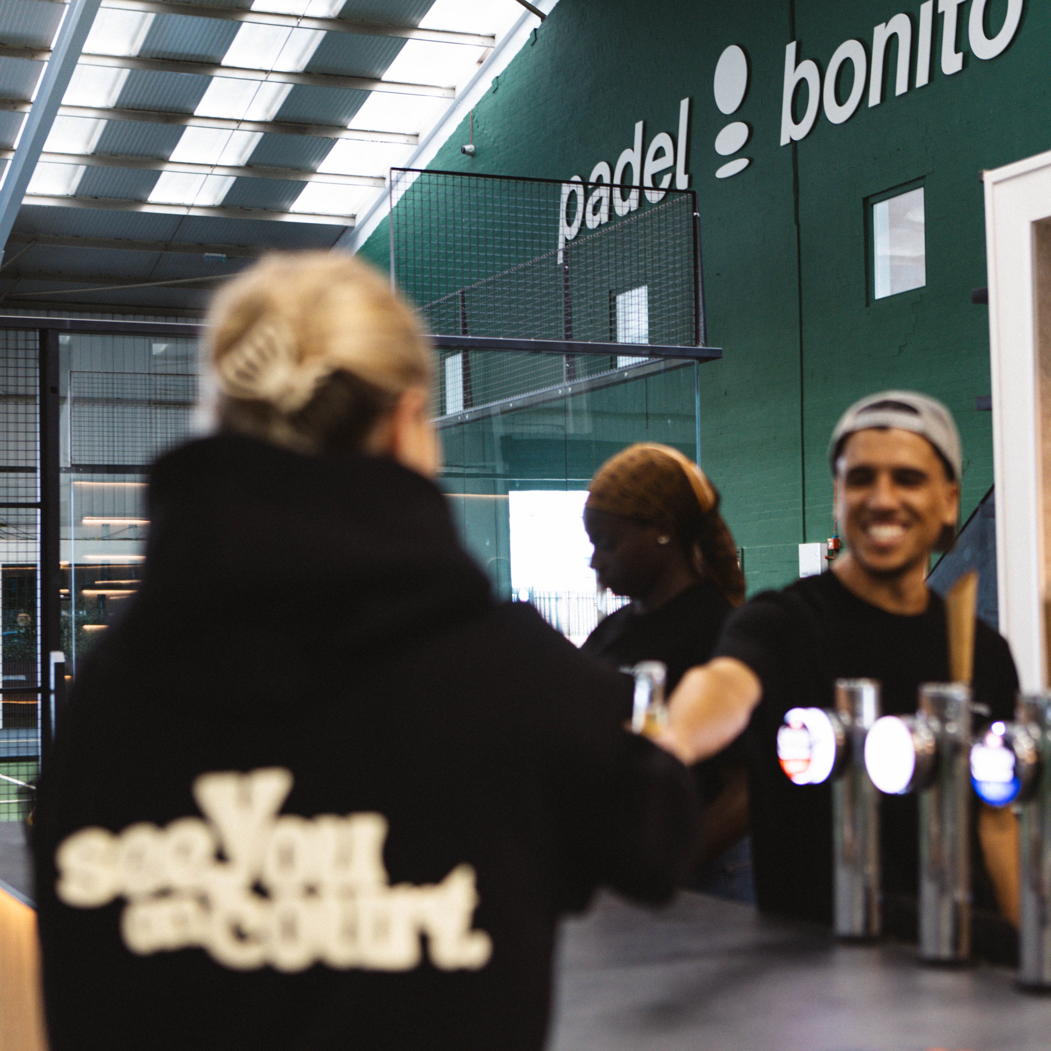 A person in a black hoodie with 'see you on court.' printed in white on the back stands in a padel venue, facing a counter with drinks.