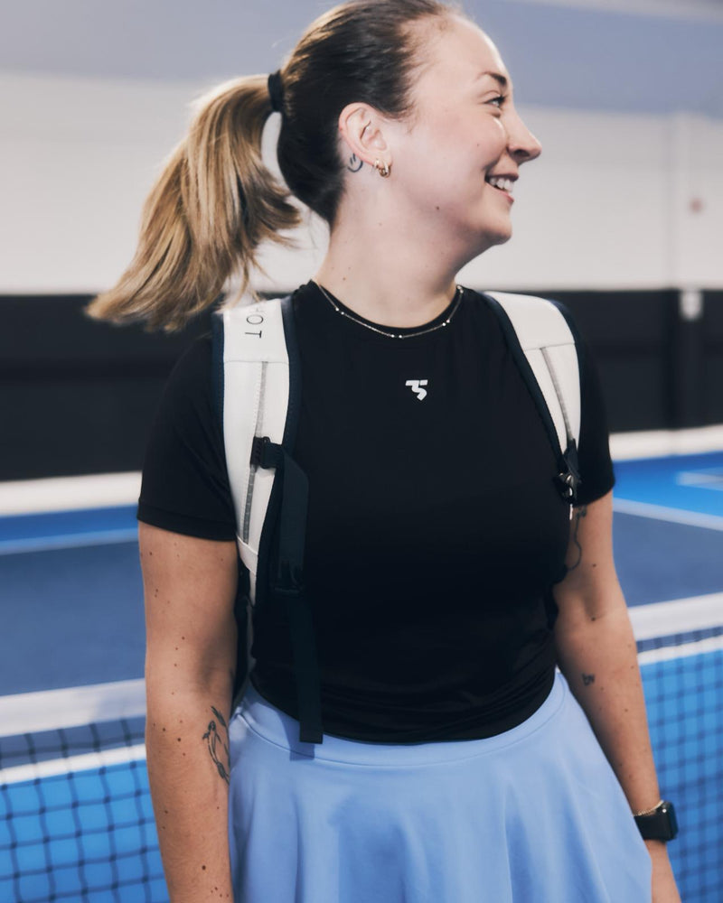 Woman on a pickleball court wearing a black sports top with a logo and a blue skirt.