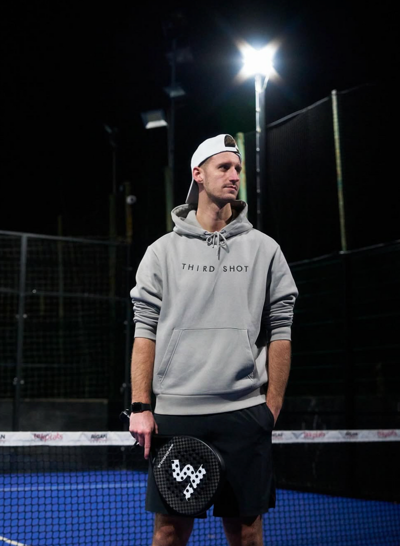 Person wearing a gray hoodie with 'Third Shot' branding on a padel court at night.