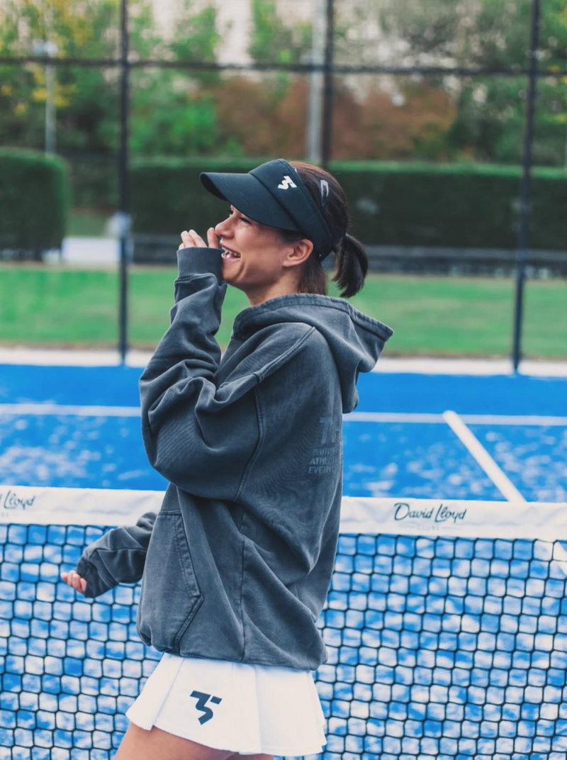 Woman on a padel court wearing a dark hoodie and cap, with a blurred background.
