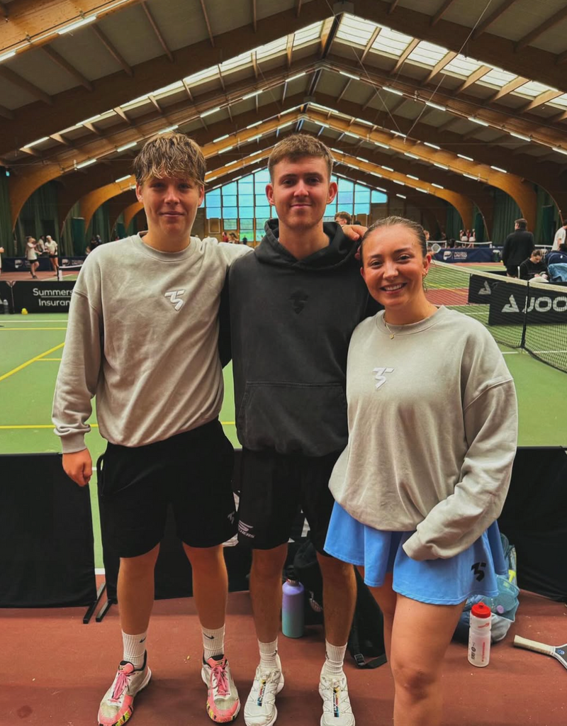 Three people posing together on a tennis court with sports equipment around.